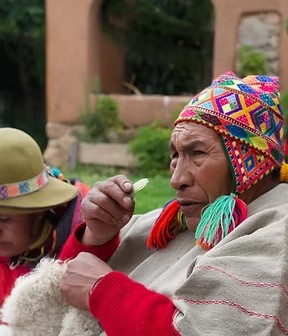 An older man in a bright knitted hat with ear flaps and a woven poncho, holding a leaf, a person in a hat beside him