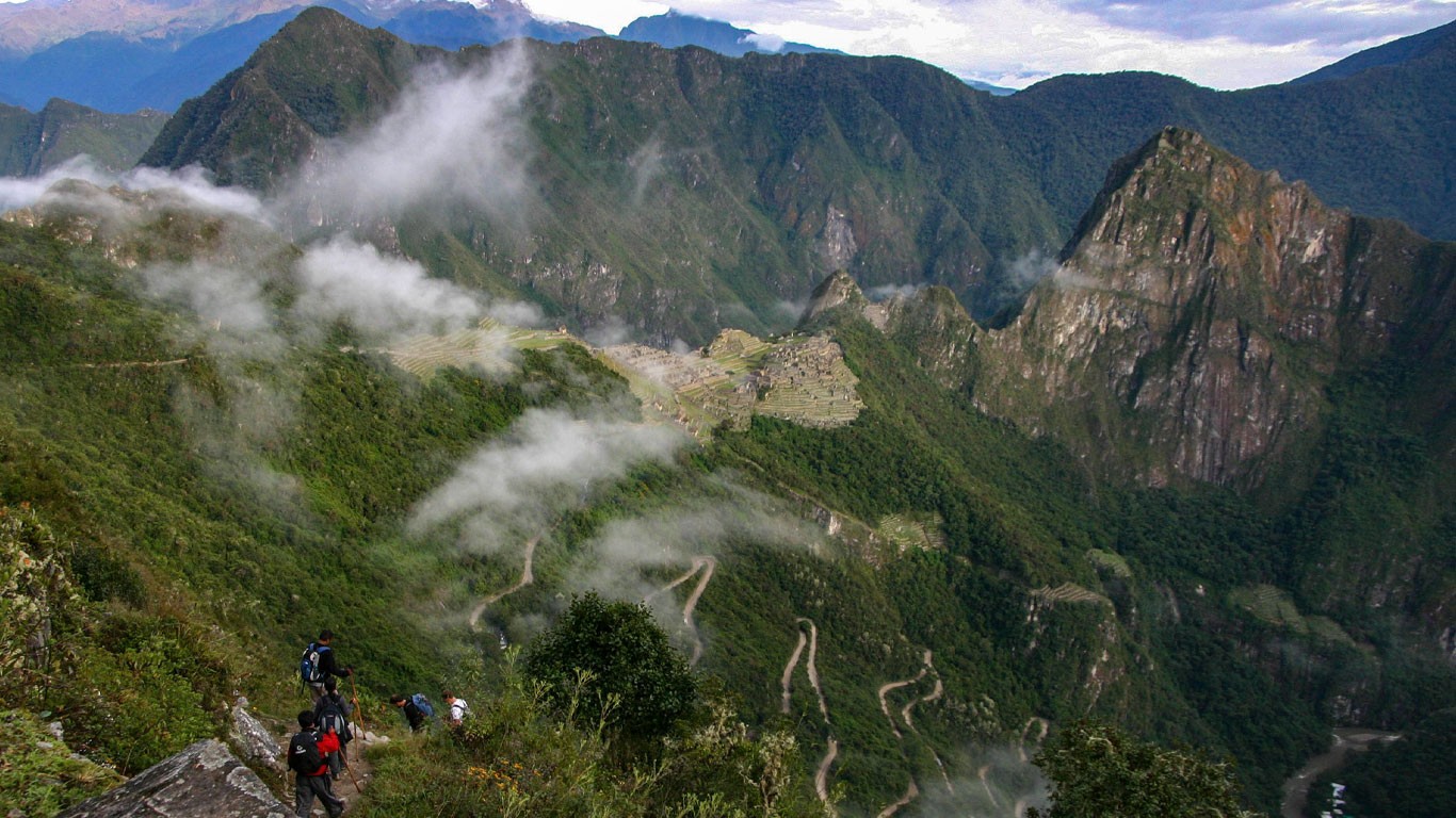 Hikers on a mountain trail above a misty green valley in Peru, a switchback road below and stone terraces on a ridge