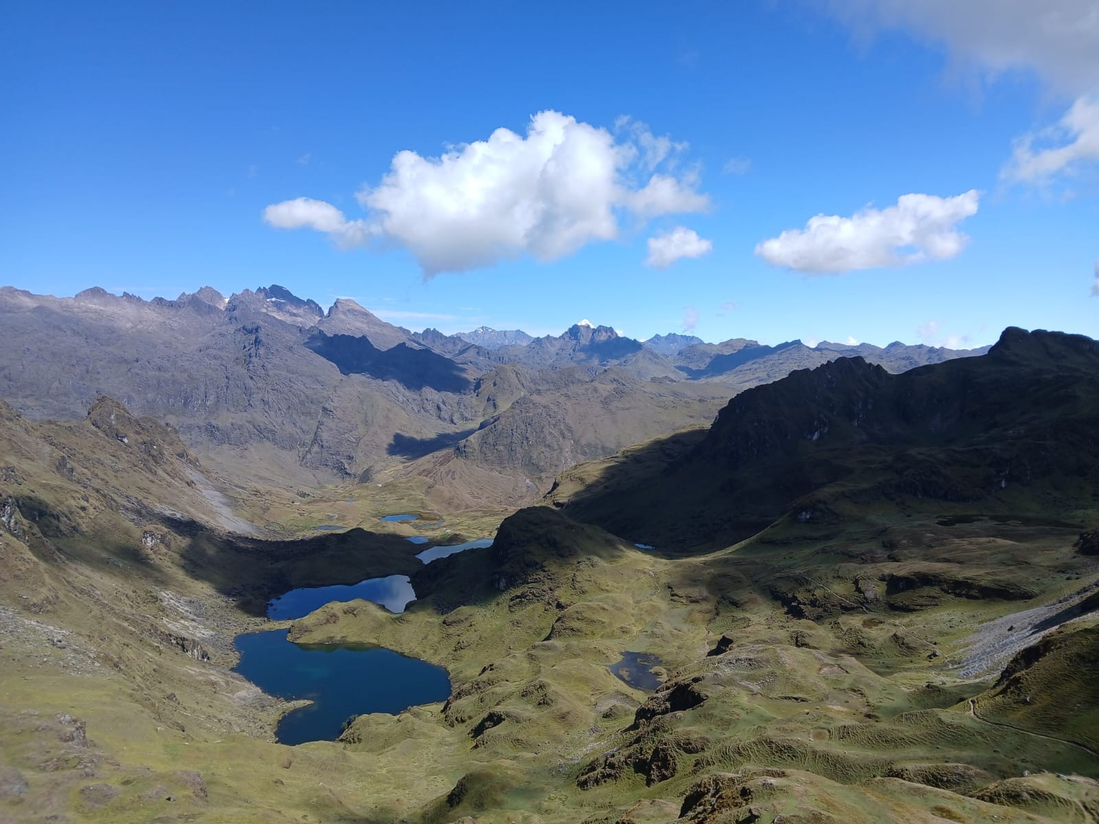 A chain of dark blue lakes in a high green valley below jagged peaks and a distant snowcapped summit