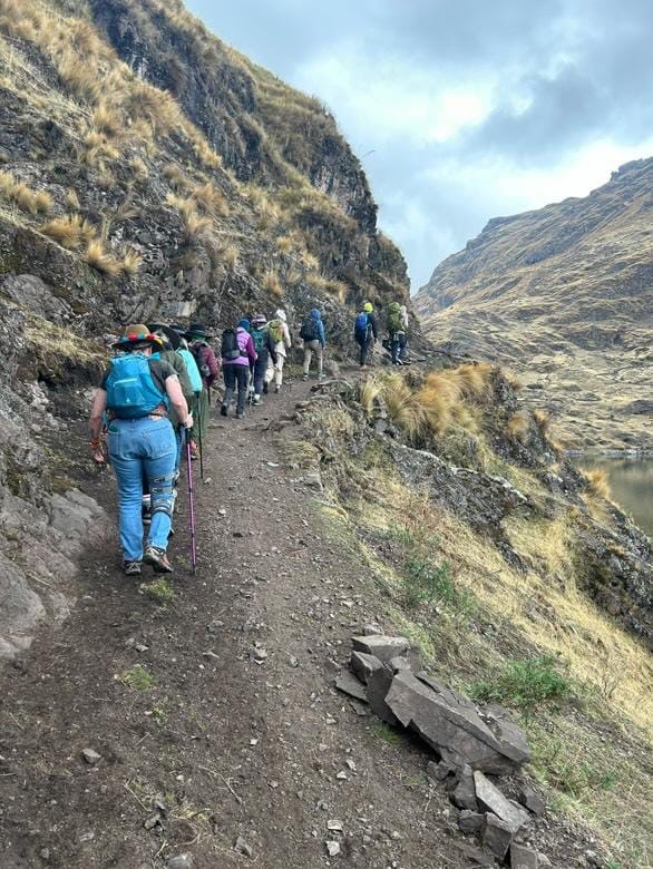 A line of hikers with backpacks and walking poles on a narrow trail along a steep grassy mountainside