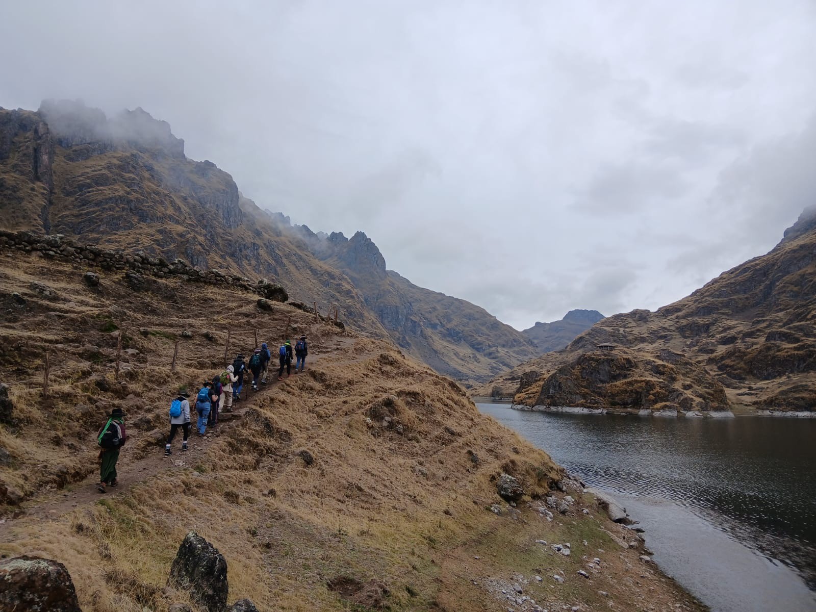 A line of hikers with backpacks on a trail along a dry golden slope beside a dark lake, mist over the peaks