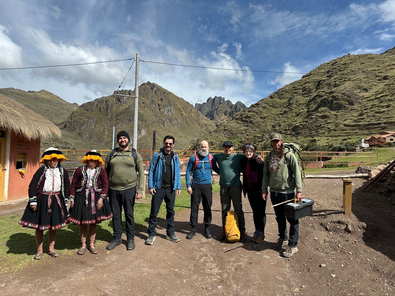 Two women in traditional dress and fringed hats stand with six hikers on a dirt road below green mountains
