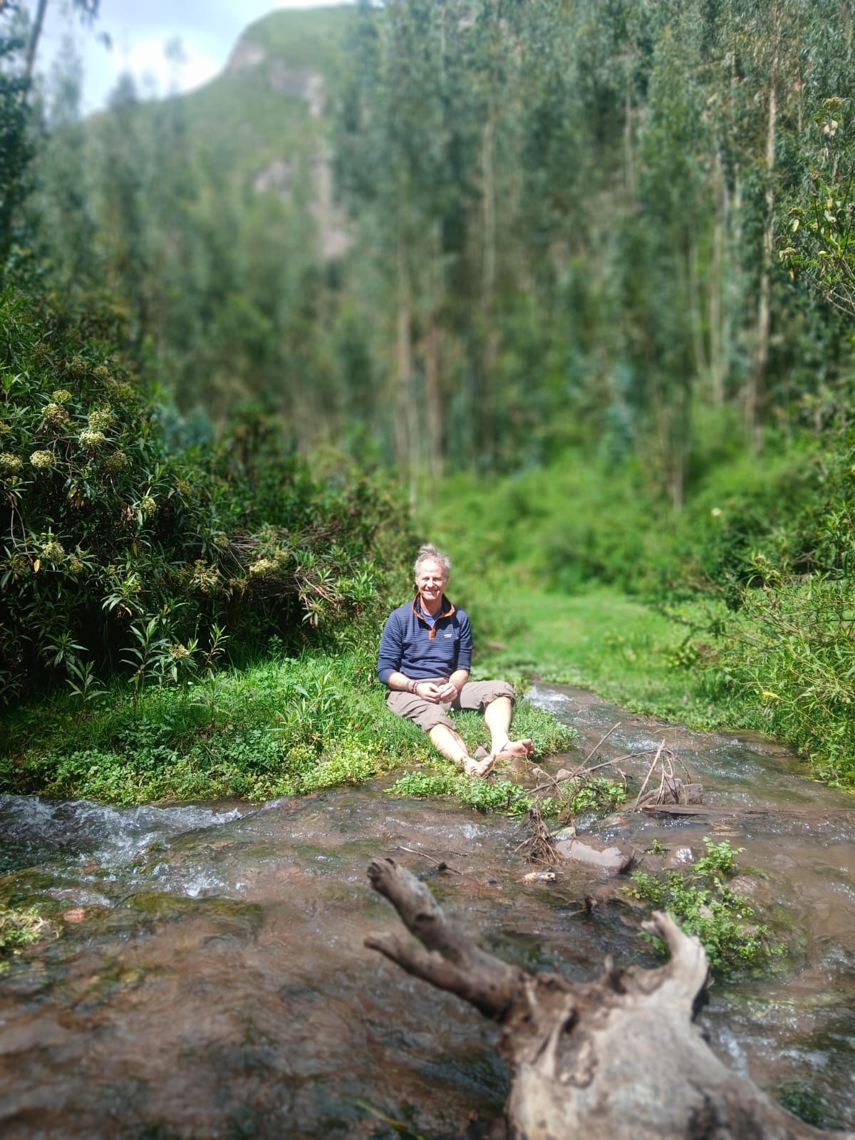 A smiling man sits barefoot on the grassy bank of a shallow stream in a forest of tall trees
