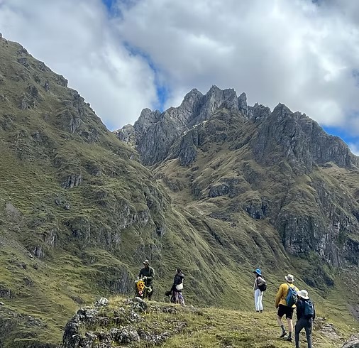 Five hikers with backpacks and sun hats on a grassy ridge below jagged rocky peaks