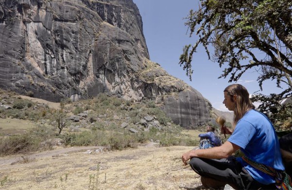 A man with long hair sits on dry grass below a towering rock face, a tree beside him