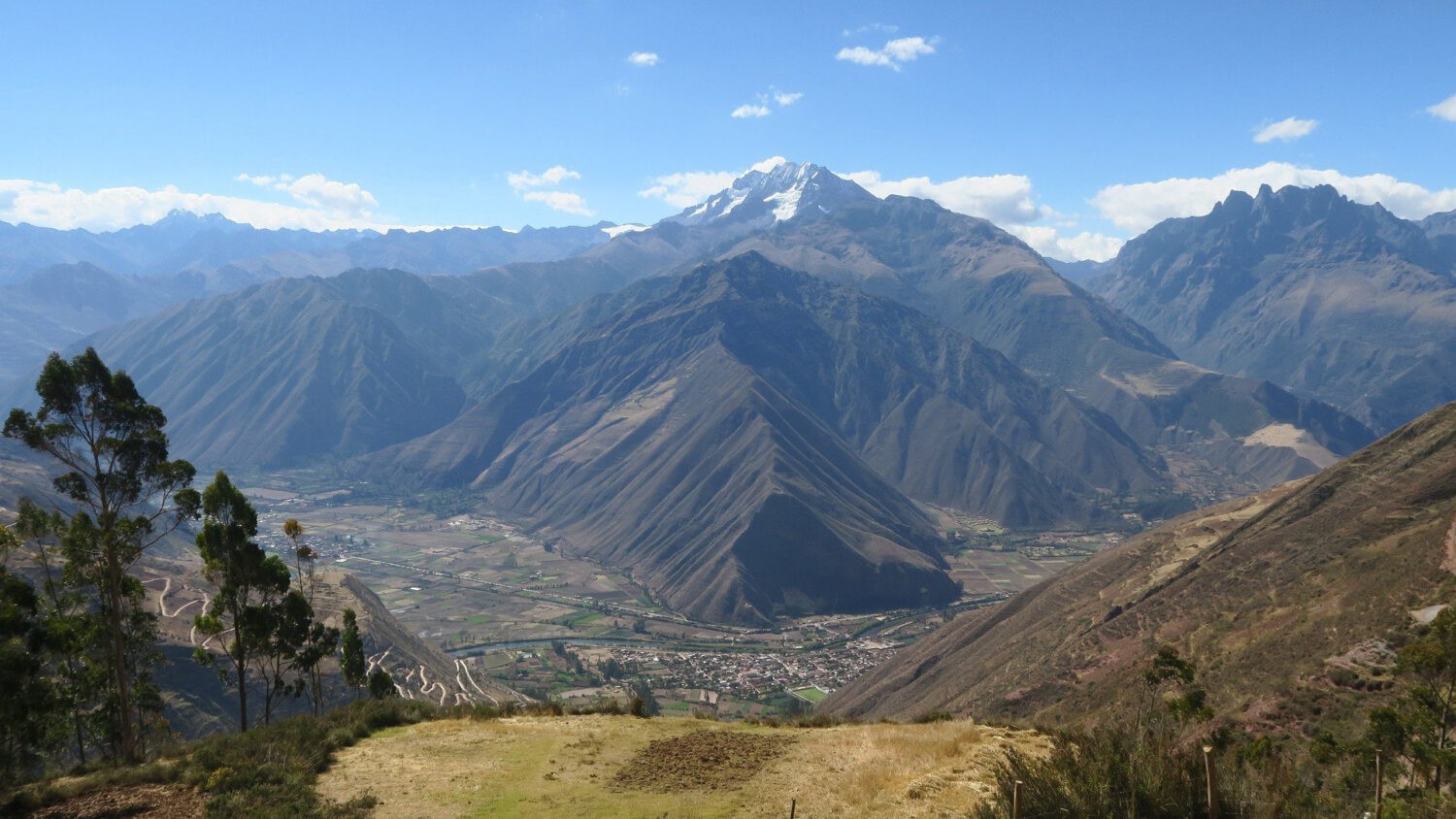 The Sacred Valley of Peru from a high grassy slope, a town and green fields far below and a snowcapped peak beyond